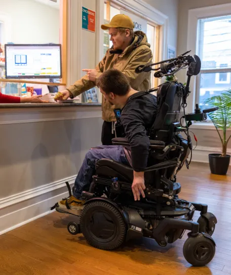 Two adults at a counter, one is in a wheelchair. Woman assisting them behind a desk.