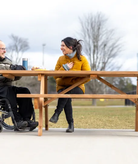Two people sitting at a picnic table outside. Both are smiling. One person is in a wheelchair.