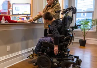 Two adults at a counter, one is in a wheelchair. Woman assisting them behind a desk.